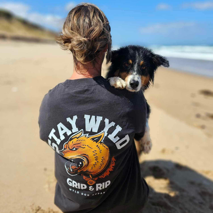 A person with their back to the camera is wearing a black t-shirt with a custom canine logo design. A dog is resting its head on the person's back, and they are standing on a beach.